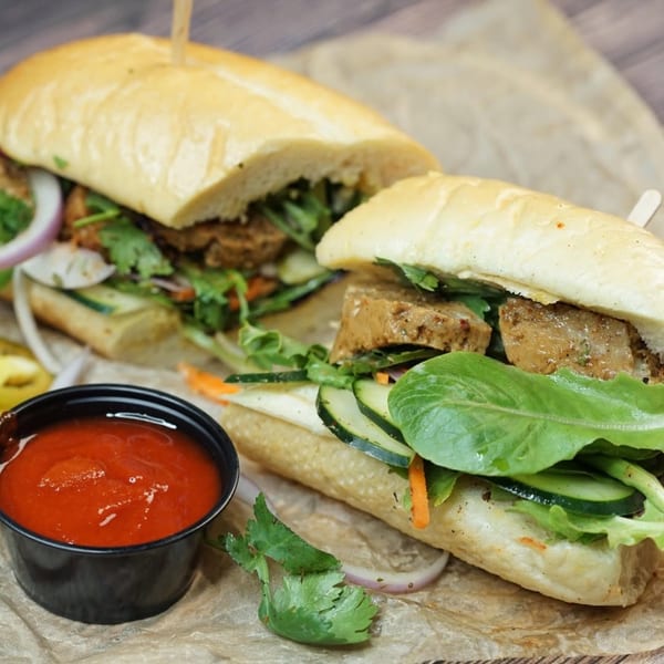 Two halves of a banh mi sandwich with meat, vegetables, and a side of red sauce.
