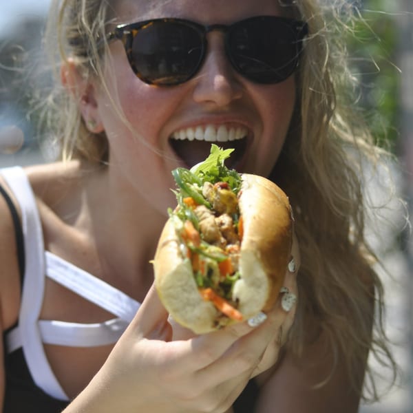 A woman in sunglasses with an open mouth, holding a sandwich filled with meat and vegetables.