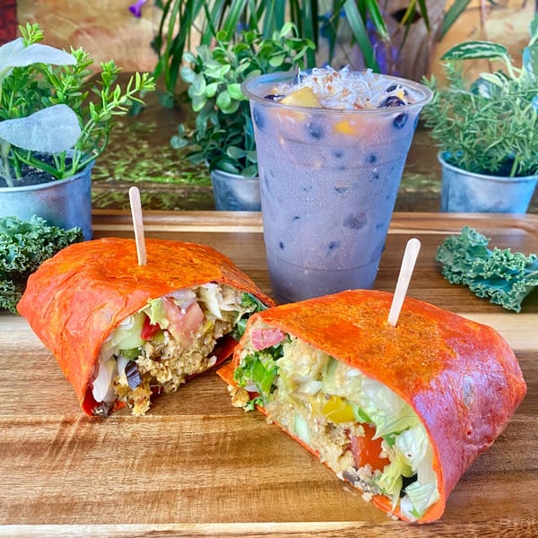 Two halves of a vibrant orange wrap filled with fresh vegetables and a purple drink on a wooden board.