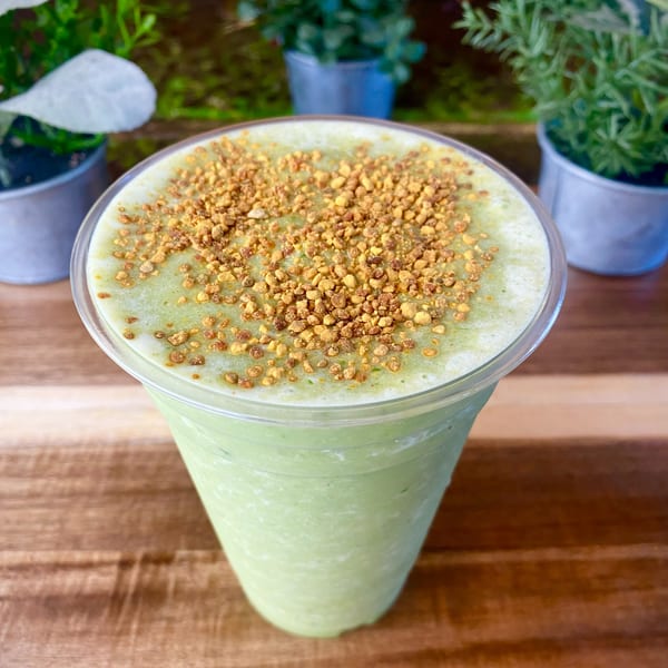 Green smoothie topped with golden bee pollen, on a wooden table with potted plants.