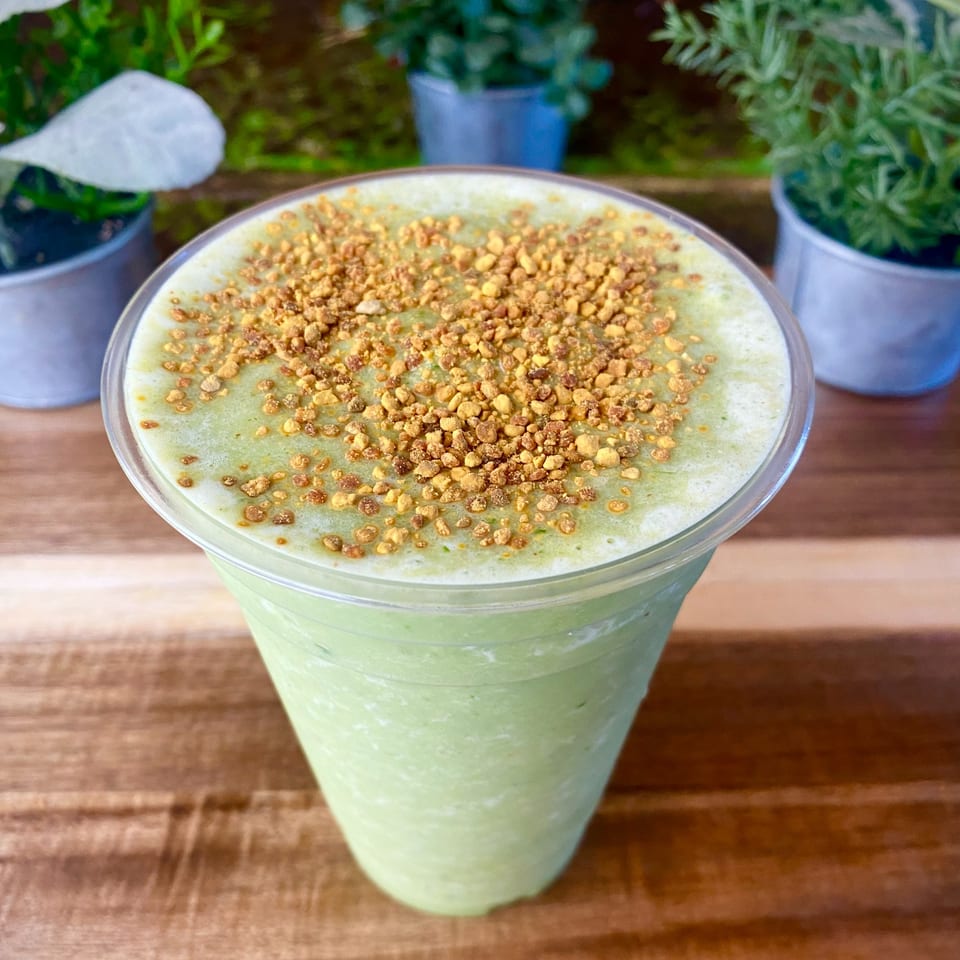 Green smoothie topped with golden bee pollen, on a wooden table with potted plants.