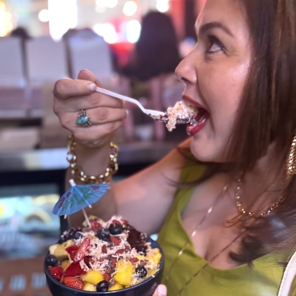 A woman in a green top eats a colorful acai bowl topped with fruit, nuts, and a tiny umbrella.
