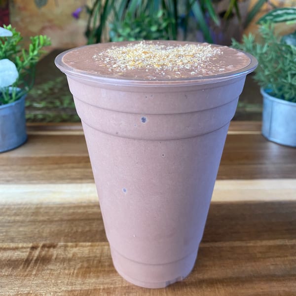 A chocolate smoothie with a sprinkle of granola on top, on a wooden table with plants.